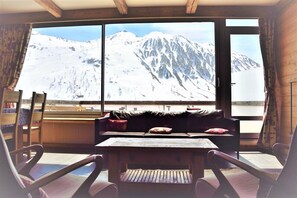 A cozy room with a large window, a wooden table, and a black sofa with red cushions.