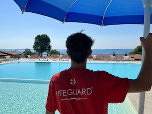 Un sauveteur se tient près d’une piscine, d’où l’on a une vue dégagée sur la mer, avec un arbre en arrière-plan.