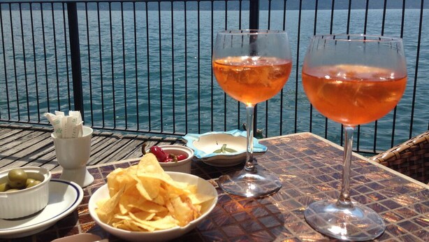 Une table avec deux verres contenant une boisson rose, un bol de chips, un petit bol d’olives et une tasse de café, le tout placé sur un balcon donnant sur une étendue d’eau.