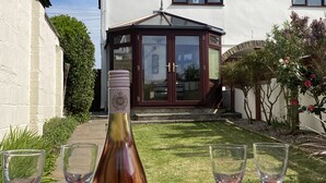 Eine Weinflasche auf einem Picknicktisch mit Gläsern, eingebettet in einen Garten mit einem Haus im Hintergrund.