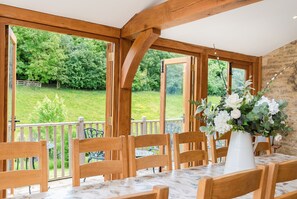A dining area with wooden chairs and a table, large windows with a view of a green landscape, and a white vase with flowers on the table. - Hollyhock House Sleeps 10, Idyllic Cotswold property. Arctic Cabin & playground. (Stonesfield)
