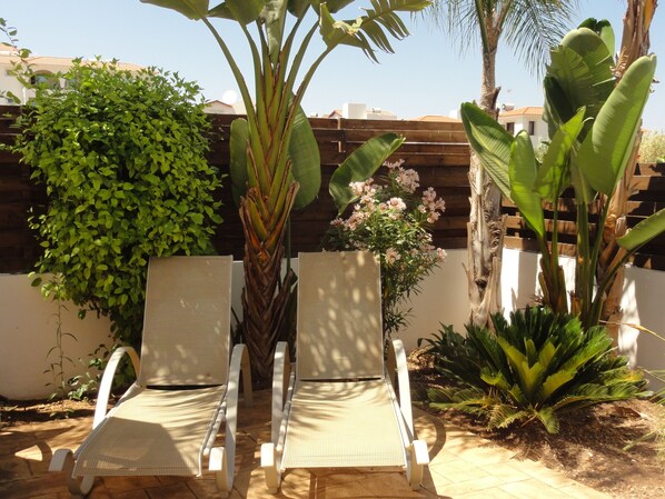 Two white lounge chairs with mesh backs, positioned in a sunlit outdoor area with a wooden fence and various tropical plants. - Stunning 3 Bed villa with pool,Wifi,Mins to the Beach & amenites (Protaras)
