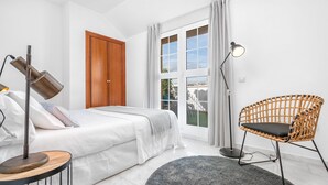 Une chambre à coucher avec un lit, une lampe sur pied, un fauteuil en osier et une fenêtre avec des rideaux.
