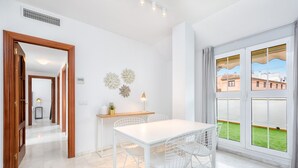 Un espace repas moderne avec une table blanche et des chaises en fil de métal, une petite table d’appoint avec une lampe, et une vue sur une pelouse verdoyante à travers de grandes fenêtres.