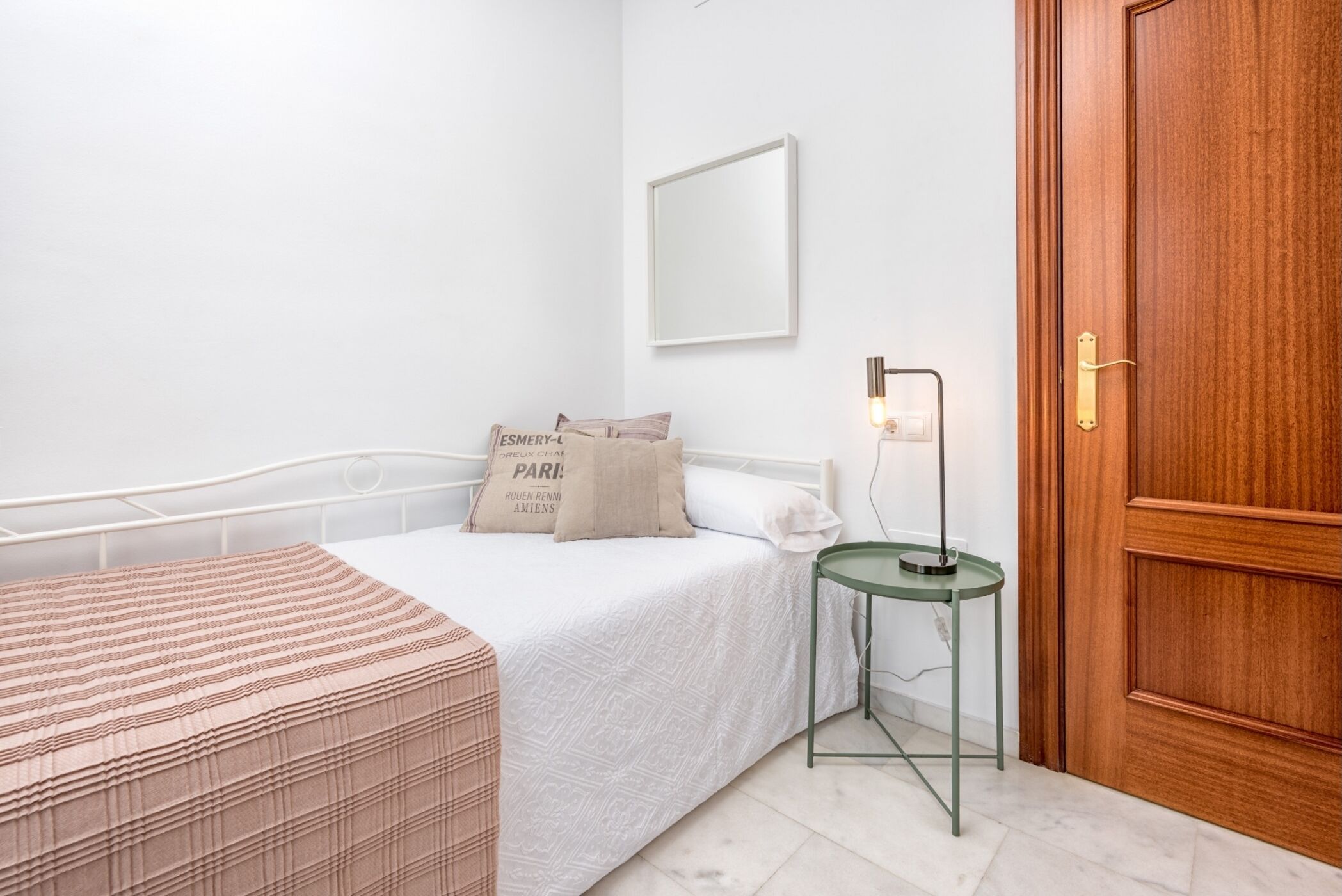 A neatly made bed with a white bedspread and two pillows, a small bedside table with a lamp, and a wooden door.