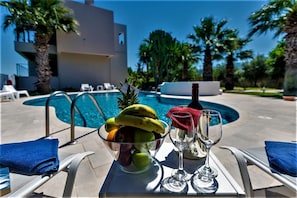 Una mesa junto a la piscina con fruteros, copas de vino y una botella, con vistas a una alberca con sillones reclinables y palmeras.