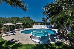 Una piscina circular con un jacuzzi, rodeada de sillas de descanso y sombrillas, con vista al mar y palmeras.