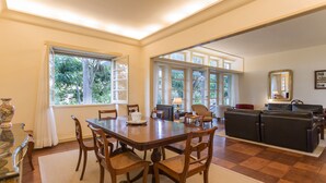 Uma sala de jantar espaçosa com mesa e cadeiras de madeira, área de cozinha com bancada e eletrodomésticos, e grandes janelas com vista para o verde.