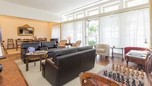 Uma sala de estar ampla com um sofá grande, uma mesa de centro e um jogo de xadrez. Há várias cadeiras e uma mesa de jantar ao fundo.