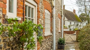 A traditional stone house with a white door and window shutters, a narrow pathway leading to a gated entrance, and a small garden area with a potted plant. - 2 Dix Cottages, Thornham, Norfolk (Thornham)