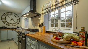 A kitchen with a large wall clock, a window with curtains, and various kitchen items on the counter. - 2 Dix Cottages, Thornham, Norfolk (Thornham)