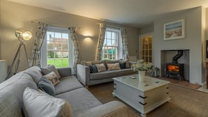 A cozy living room with a sofa set, a white coffee table, a fireplace, and large windows with curtains. - 2 Dix Cottages, Thornham, Norfolk (Thornham)