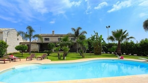 Piscina com espreguiçadeiras, palmeiras e um edifício de pedra ao fundo.