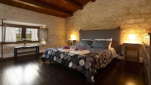 Quarto com parede de pedra, vigas de madeira no teto, cama com colcha estampada, duas luminárias de cabeceira e mesa de cabeceira com abajur.