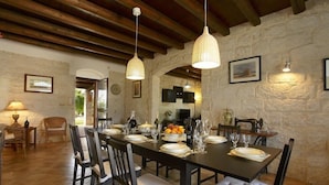 Uma sala de jantar com parede de pedra, vigas de madeira no teto e uma mesa comprida posta para seis lugares, com cadeiras.