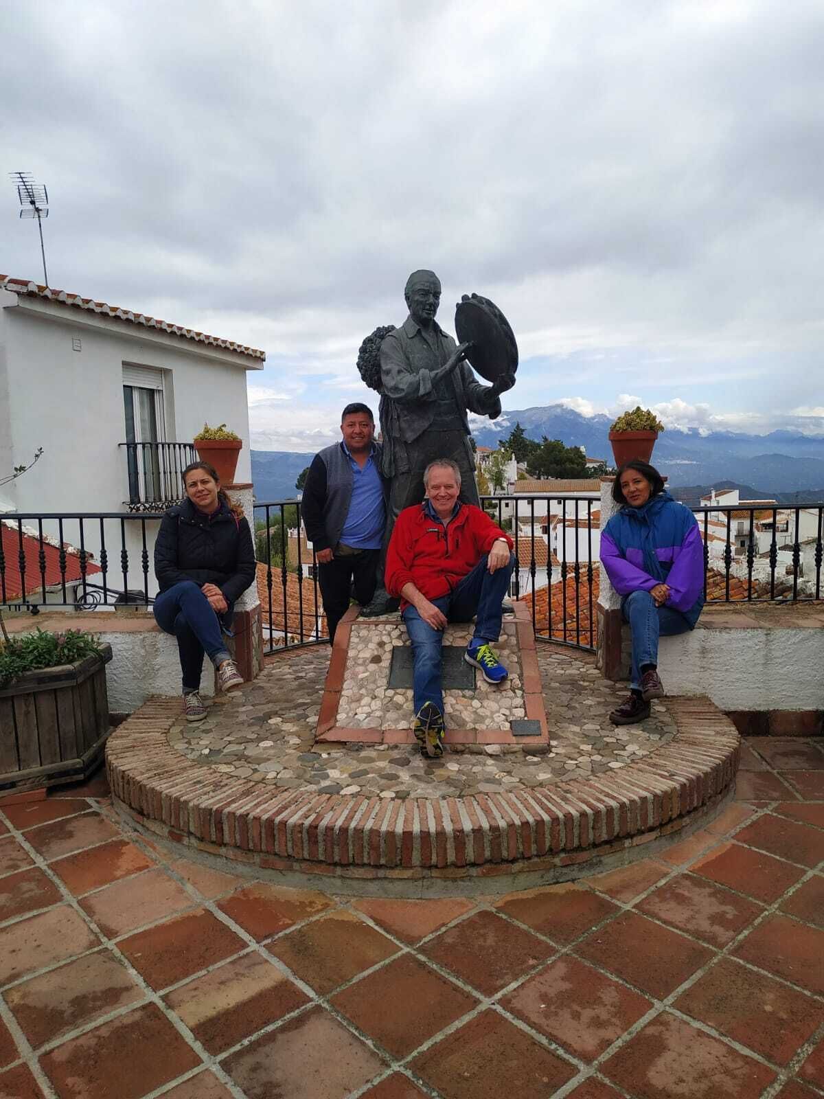 A group of people sitting on a stone bench with a statue in the center, surrounded by a metal fence and potted plants.