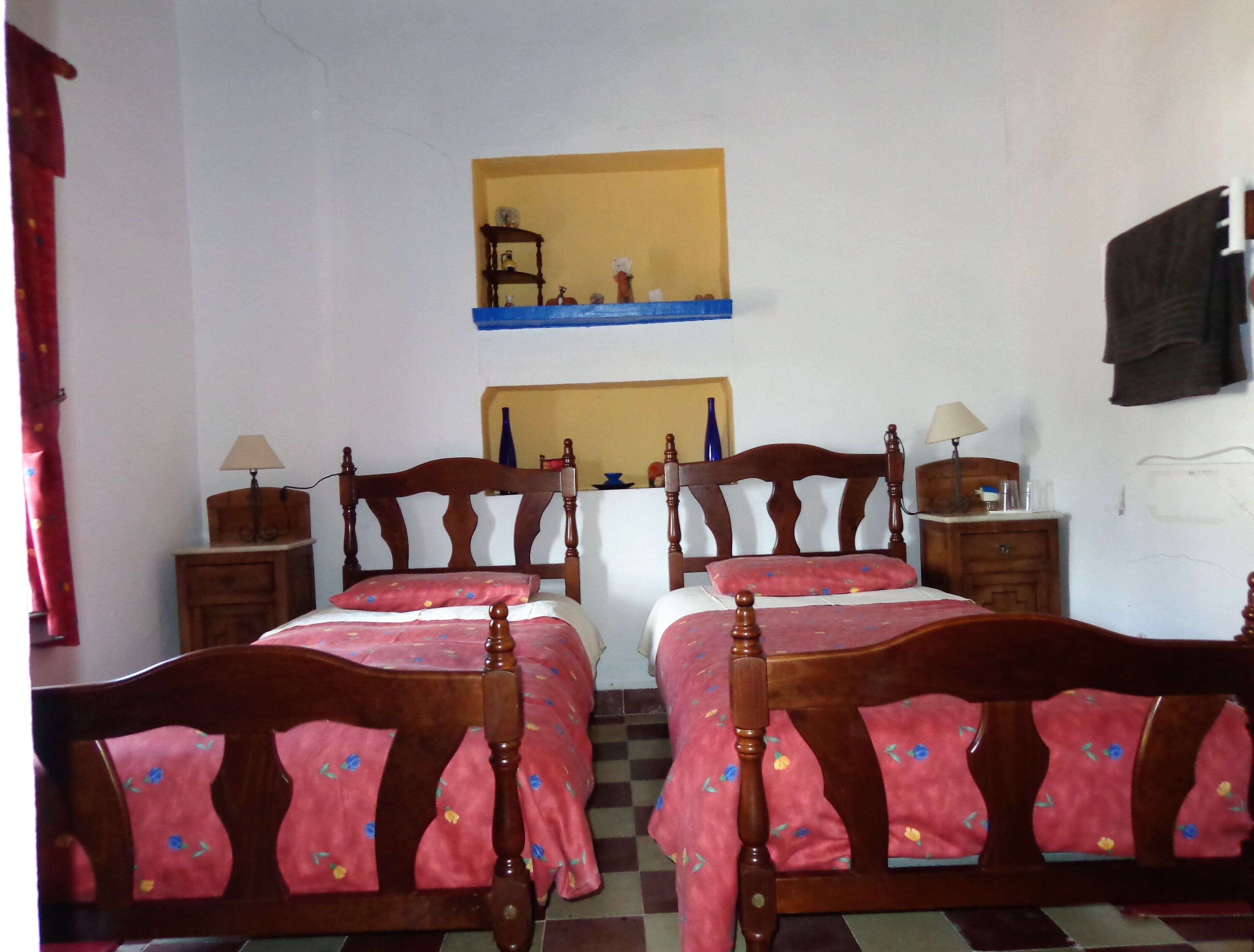 A room with two wooden beds with floral bedding, a small table with a lamp, a mirror on the wall, and a red curtain.