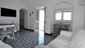 A modern living room with a white sofa, a dining area with a table and chairs, and a kitchen area with a window and cabinets. - Amalfi Coast - Il Monaco, Three Bedrooms villa with equipped platform on the sea (Ravello)