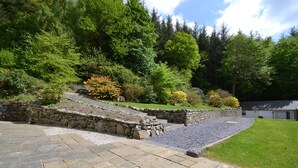 A stone patio leading to a stone wall, surrounded by lush greenery and trees. - Spacious, detached & private Lakeland hideaway near Cark / Cartmel (Cumbria)