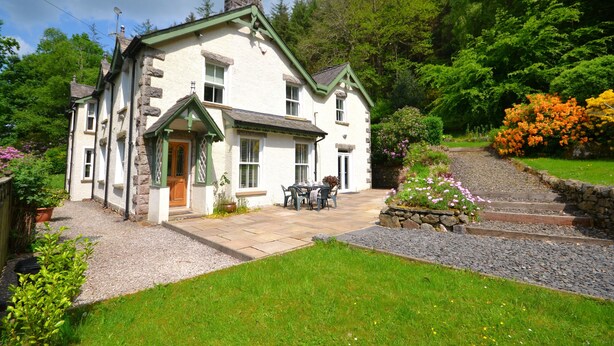 A white house with a stone facade, a front porch, and a garden with flowers and a gravel path. - Spacious, detached & private Lakeland hideaway near Cark / Cartmel (Cumbria)