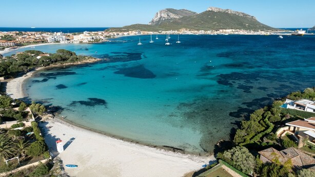 Vue aérienne d’une zone côtière avec des eaux turquoise limpides, une plage de sable et un paysage montagneux en arrière-plan.