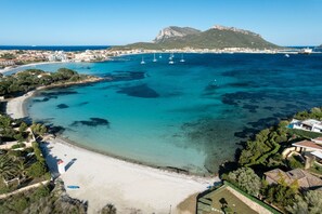 An aerial view of a coastal area with clear turquoise waters, a sandy beach, and a mountainous backdrop. - Villaggio Perlacea - PERLACEA 19 (Golfo Aranci)