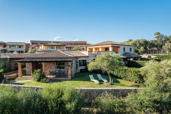 A residential area with multiple houses featuring Mediterranean architecture, stone walls, and a well-maintained garden with a lawn and trees. - Villaggio Perlacea - PERLACEA 19 (Golfo Aranci)