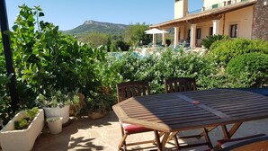 An outdoor patio with a wooden table and chairs, surrounded by potted plants and a view of a mountain and pool in the background. - Country villa with pool, private garden and barbecue near the beach OFFER June (Manacor)