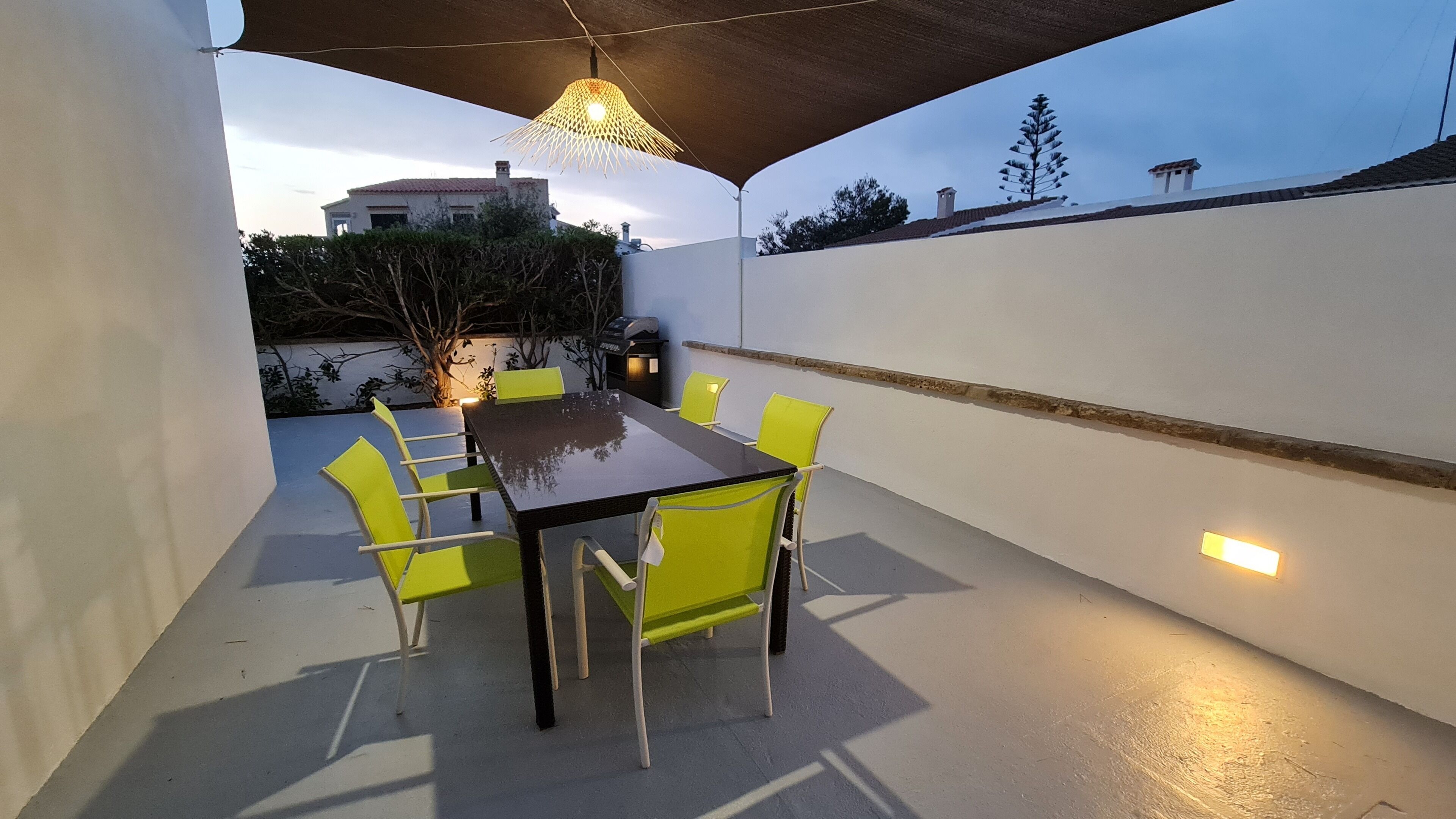 Un patio exterior con una mesa y sillas, un colgador de luces y vista a un edificio al fondo.