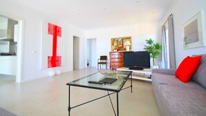 Een moderne woonkamer met een glazen salontafel, een grijze bank met een rood kussen, een flatscreen televisie en een abstract kunstwerk in rood op de muur.