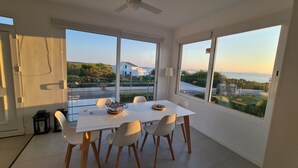 Een moderne eethoek met een witte tafel en stoelen, grote ramen met uitzicht op zee en groen, en een plafondventilator.