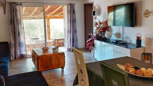 A living room with a sofa, a coffee table, a dining table with chairs, a TV mounted on the wall, and a view through the glass door. - Santa Giulia Beach & Charming stone house (porto vecchio)