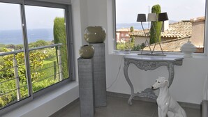 Une pièce dotée d’une grande fenêtre donnant sur un jardin et la mer, d’une statue de chien blanc et d’une table agrémentée d’objets décoratifs.