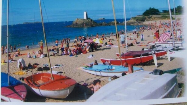 Una playa con varios barcos pequeños, un faro y gente disfrutando del día soleado.