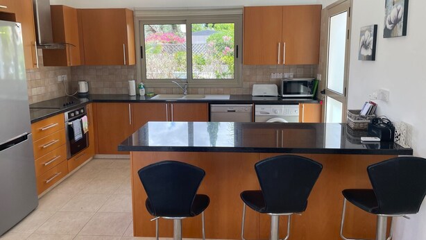 A modern kitchen with wooden cabinets, a central island with bar stools, stainless steel appliances, and a window with a view of greenery.