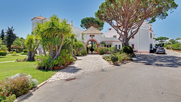 A large, two-story house with a central archway, surrounded by lush greenery and a paved driveway. - Freshly renovated with AirCo, ground floor apt with terrace, garden and pool (Vilamoura)