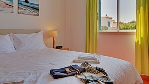 A hotel room with a bed, a nightstand with a lamp, a window with curtains, and two beach-themed wall art pieces. - Freshly renovated with AirCo, ground floor apt with terrace, garden and pool (Vilamoura)