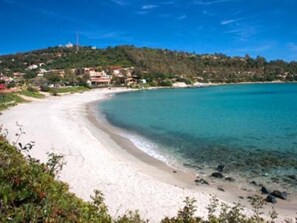 Una playa con agua turquesa cristalina, arena blanca y un paisaje montañoso con edificios al fondo.