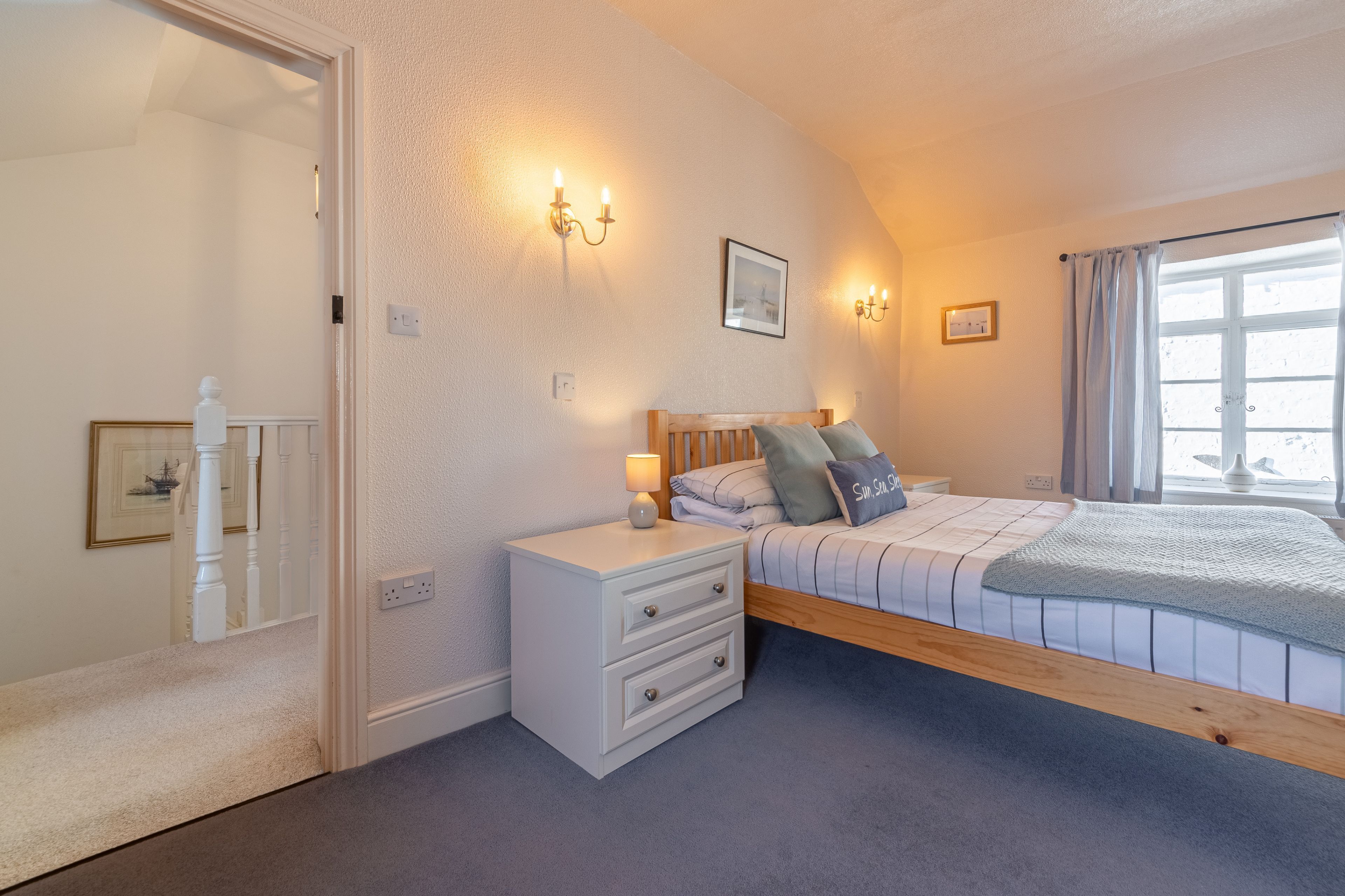 Un dormitorio con una cama de madera, una mesita de noche con cajones, una lámpara de pared y una ventana con cortinas.