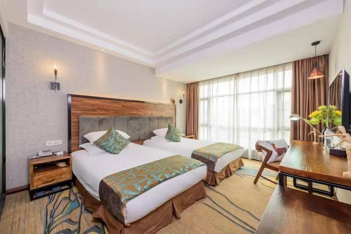 Una habitación de hotel con dos camas, un cabecero de madera, una mesa de comedor y un ventanal con cortinas.