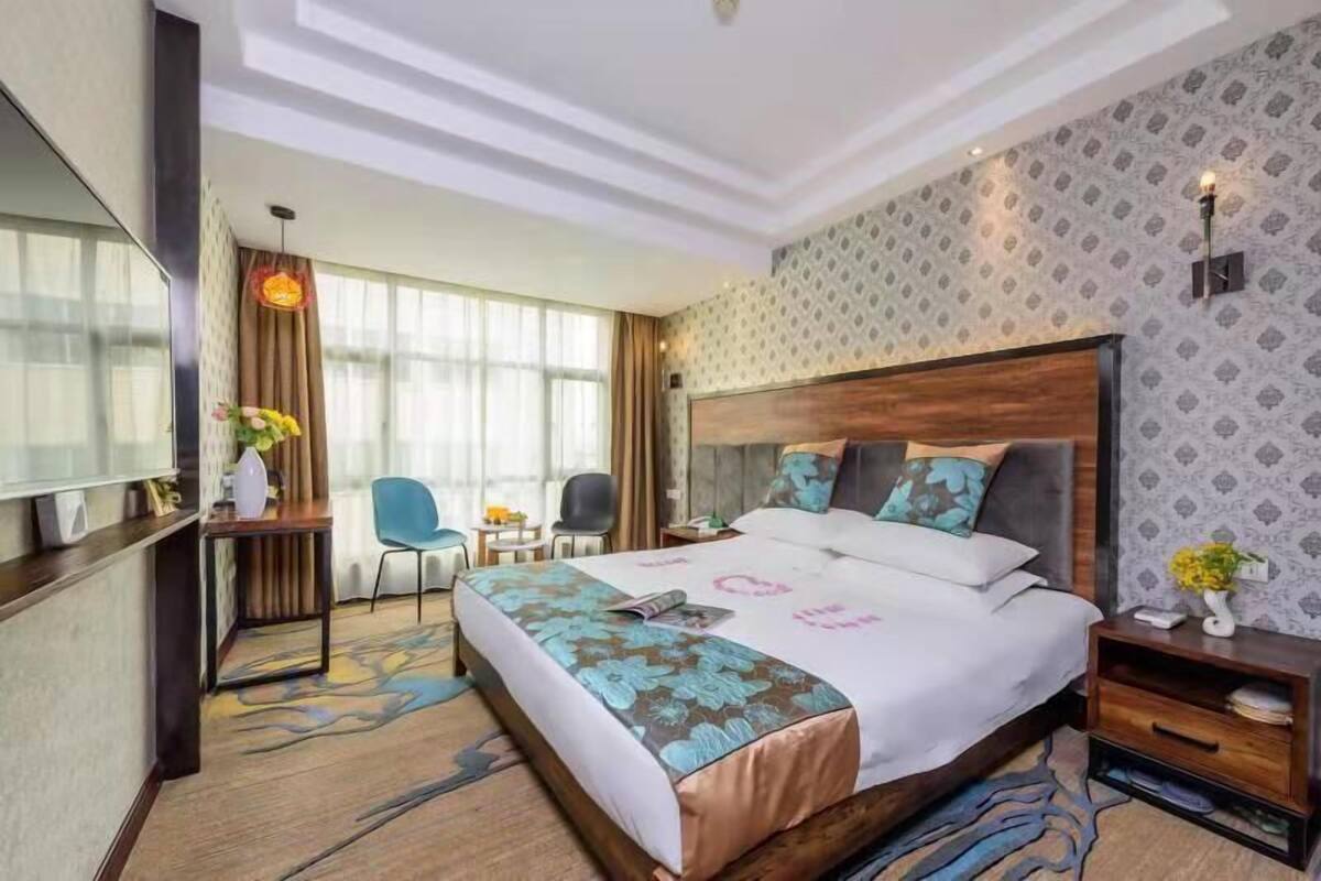 Habitación de hotel con una cama grande, un escritorio con silla, una mesita de noche con lámpara y un ventanal con cortinas.