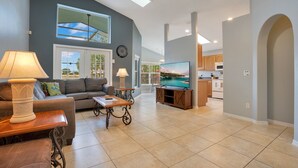 A spacious living area with a sofa, coffee table, and TV, connected to a kitchen with wooden cabinets and a large window. - *Tinkerbells Castle 2*  Private Pool & Spa Overlooking Lake & Disney Fire Works (kissimmee)