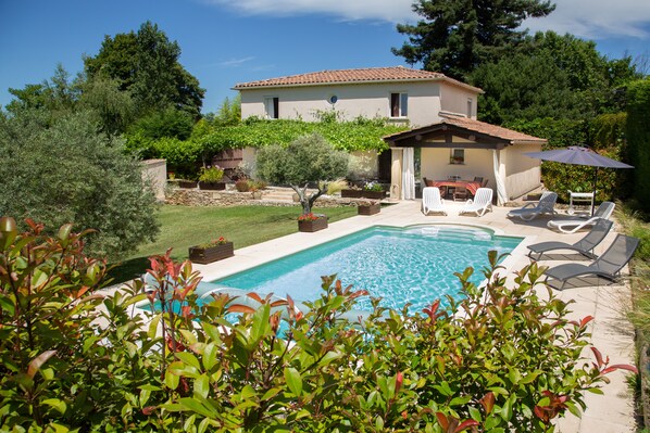 Zona junto a la piscina con una casa, mobiliario exterior y exuberante vegetación.