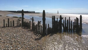 Une plage de galets, parsemée de poteaux en bois, et un phare visible au loin.