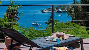 Un balcón con vistas al mar, una silla de descanso, una mesa con un sombrero y una bebida, y una planta en maceta.