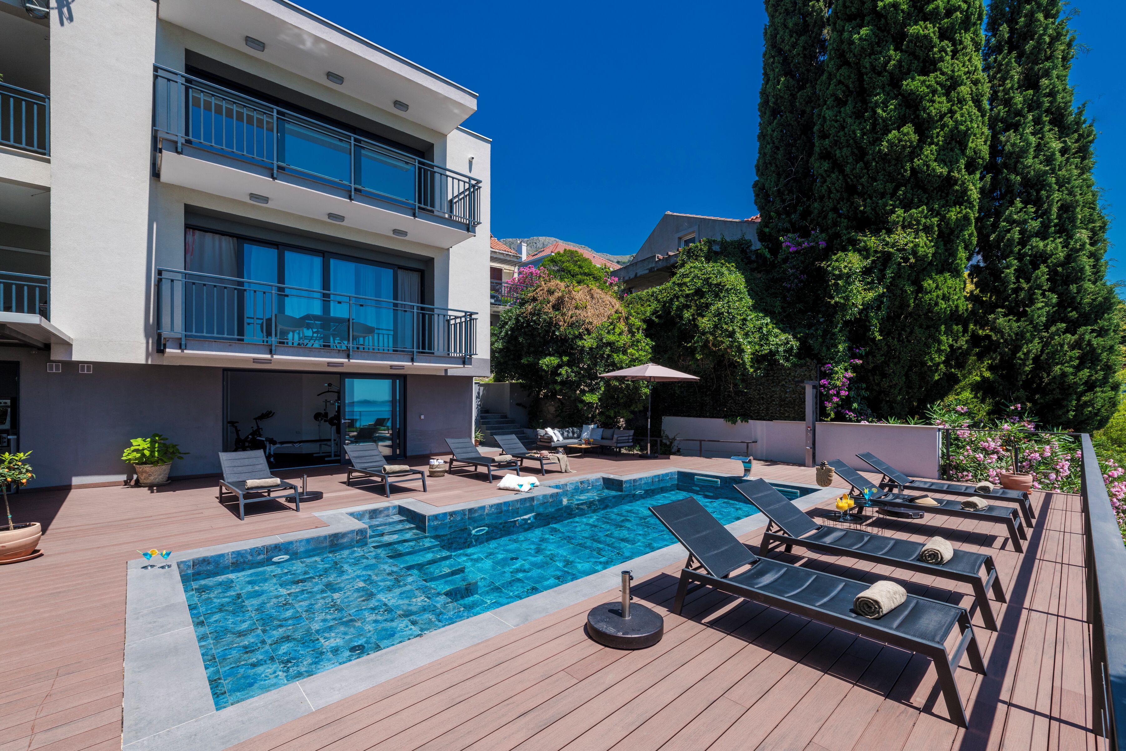 A modern building with a rooftop pool, sun loungers, and a large tree.