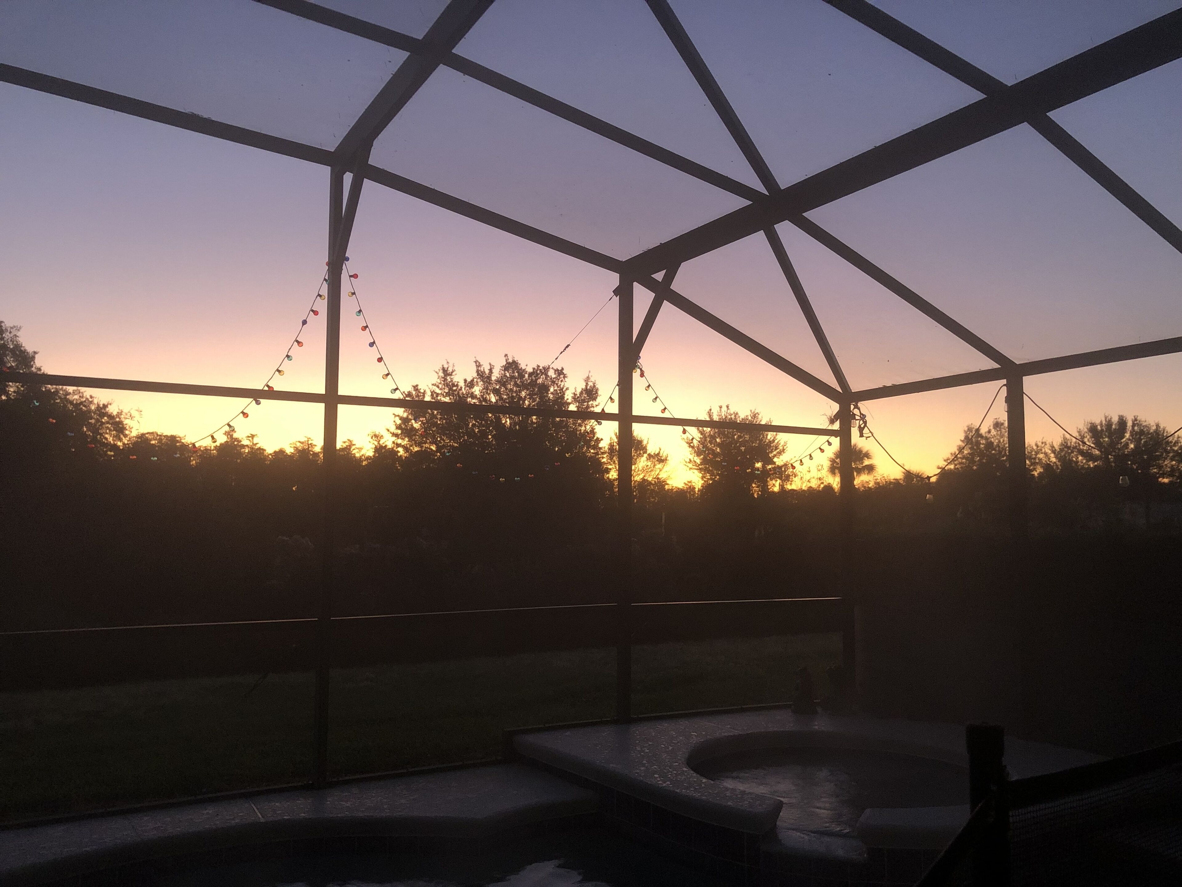 A covered outdoor area with a hot tub, metal framework, and string lights against a sunset sky.