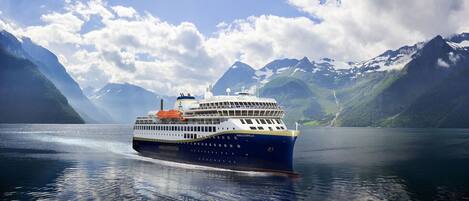 A cruise ship named "HAVILA KYSTRUTEN" sailing on a calm lake with mountainous landscape in the background.