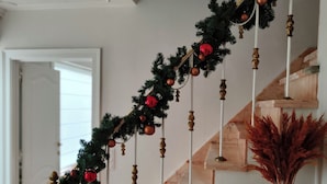 Un escalier orné d’une guirlande de décorations rouges et dorées, un vase contenant des plantes séchées et une étagère en bois avec des livres et un panier.