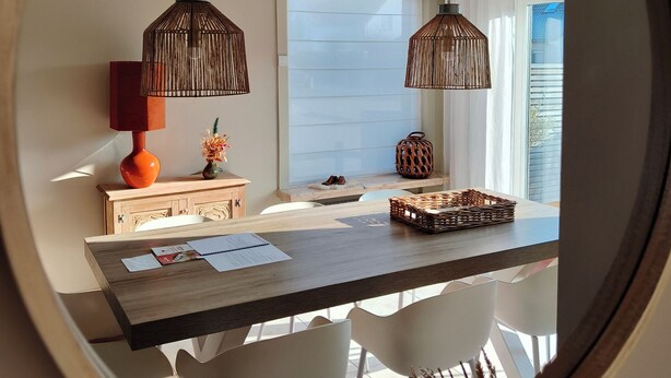 Un coin repas agrémenté d’un grand miroir rond qui reflète une cuisine moderne, avec des luminaires suspendus, une table en bois et des chaises blanches.